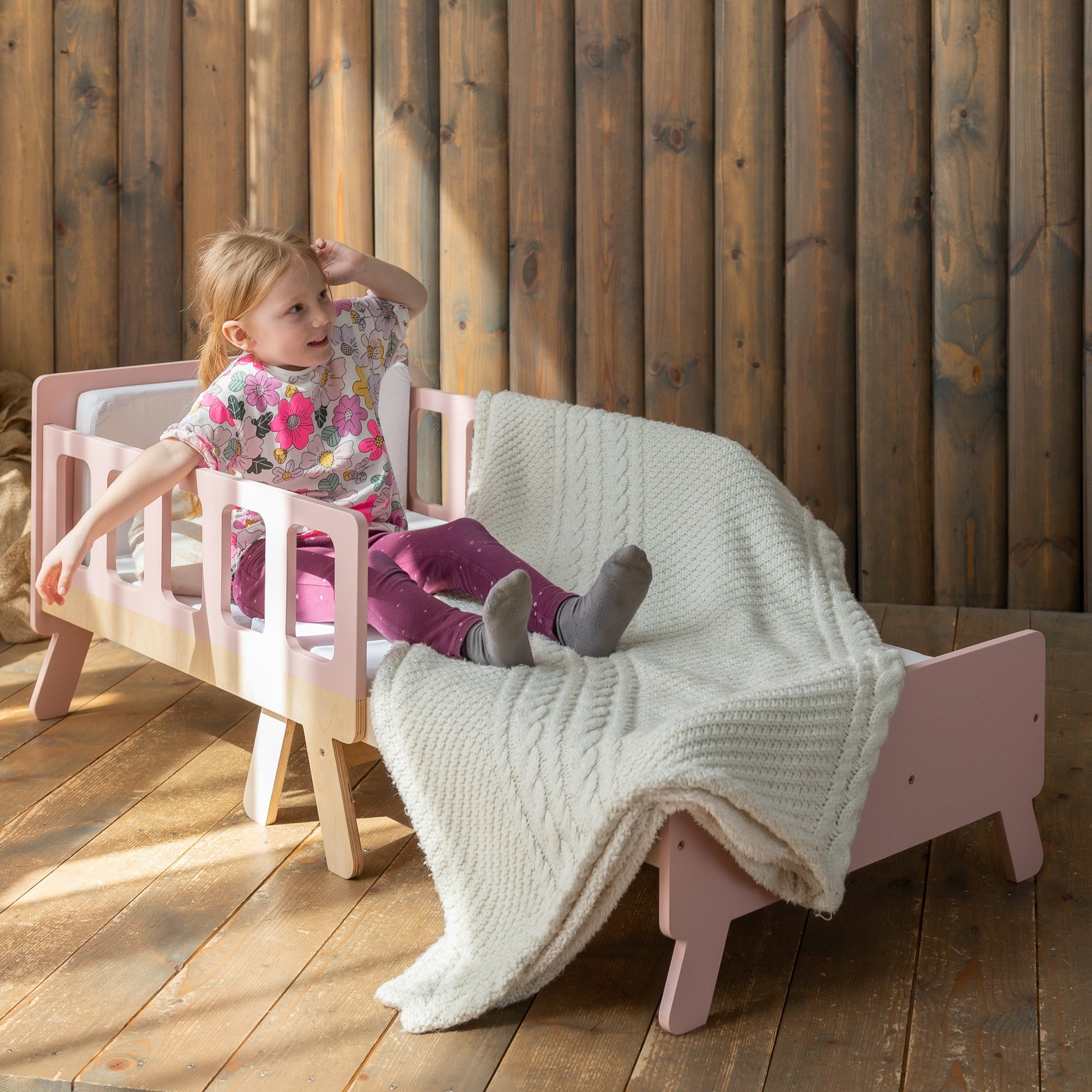 Child sitting on a pink chair with a white blanket in a wooden room.