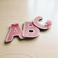 Pink checkered letters 'A', 'B', and 'C' on a wooden surface